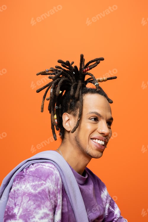 Preview: portrait of positive multiracial man with dreadlocks and piercing looking away isolated on orange