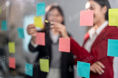 Preview: Two businesswomen planning project using adhesive notes on glass wall in office