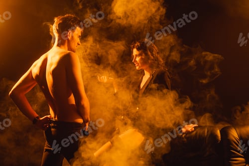 Preview: curly woman in jacket looking at shirtless man standing with hand on hip on black with smoke