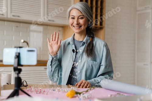 Preview: Smiling grey woman gesturing while taking selfie footage on cellphone