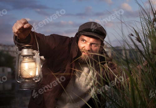 Preview: a man with a beard in vintage clothes and with a kerosene lamp in his hand