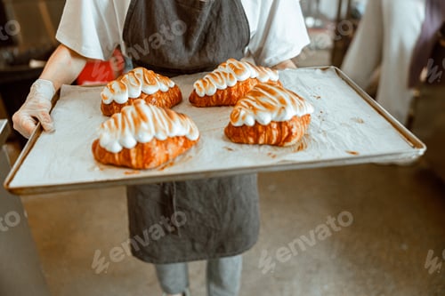 Preview: Woman in long apron holds large tray with fresh decorated croissants in craft bakery shop