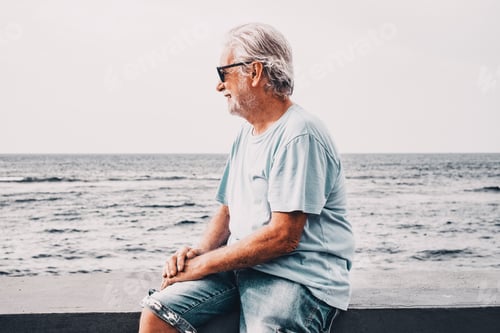 Preview: Portrait of relaxed satisfied senior bearded man sitting on sea beach at sunrise looking at horizon