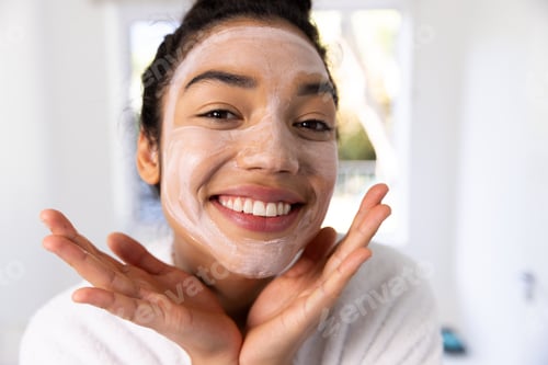 Preview: Portrait of happy biracial woman in bathrobe with face mask in sunny bathroom