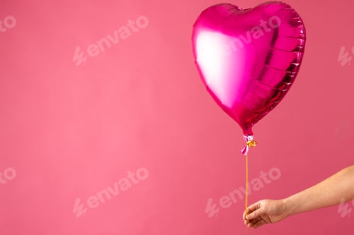 Preview: Hand holding shiny pink heart shaped balloon floating on pink background with copy space