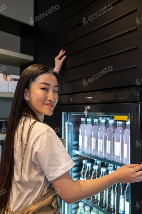 Preview: Waist-up portrait of a pleased bartender leaning on the glass door of the refrigerated display