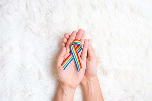 Preview: hand showing LGBTQ Rainbow ribbon on white bed background.