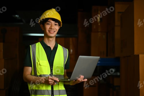 Preview: Smiling male manager wearing hardhat and reflective jacket standing between shelves.