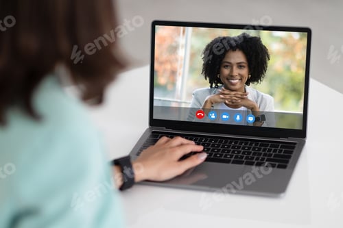 Preview: Young caucasian businesswoman in suit at table with laptop with happy black lady on screen