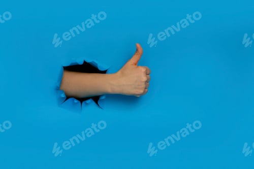 Preview: Female hand gestures ok approval sign keeps thumb up recommends something breaks through blue paper
