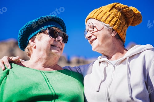 Preview: Happy couple of elderly women wearing glasses and winter hats hugging in outdoor mountain