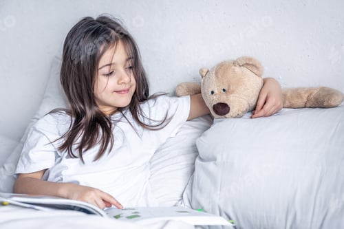 Preview: Little girl reads a book with a teddy bear in bed in the morning.