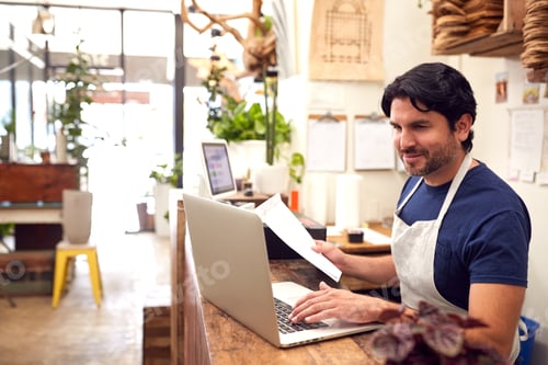 Preview: Male Sales Assistant Working On Laptop Behind Sales Desk Of Florists Store
