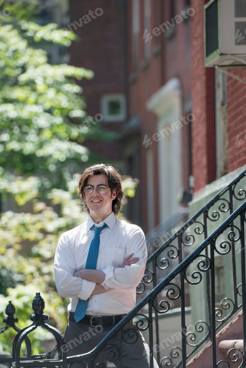 Preview: A young man in a white shirt and blue tie, standing outside a townhouse, on the steps.