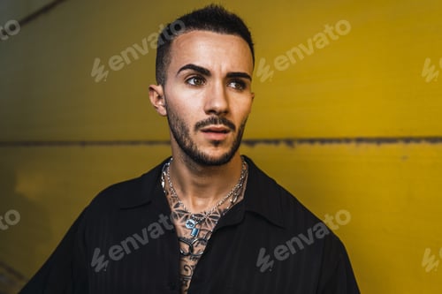 Preview: Portrait of a young hot tattooed man wearing a black shirt standing against a bright yellow wall