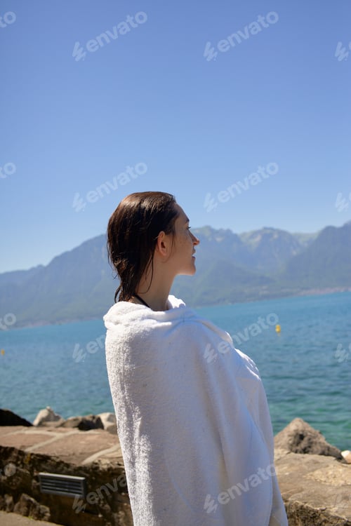 Preview: A person wrapped in a white towel stands by a lake gazing at mountains under a clear blue sky.