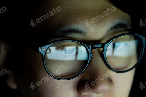 Preview: Young Asian man in glasses watching videos and surfing internet on technology device in the dark