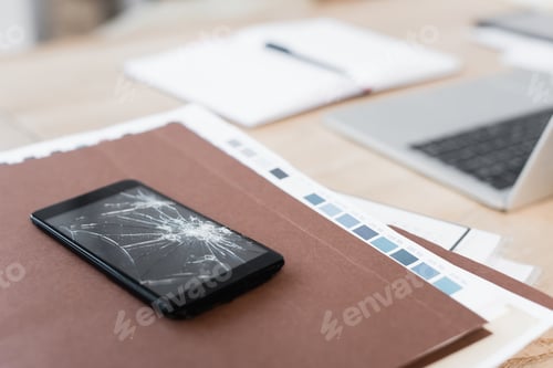 Preview: Close up view of smashed smartphone on pile of paperwork with blurred workplace on background