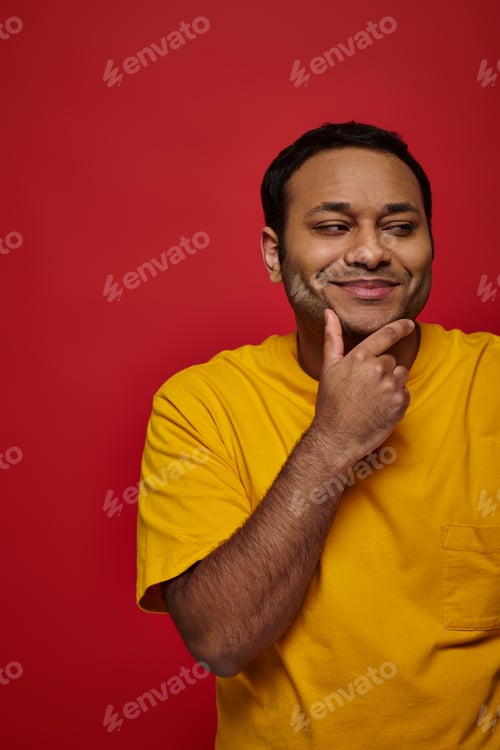Preview: pleased indian man in yellow t-shirt touching chin and looking away on red background, thoughtful