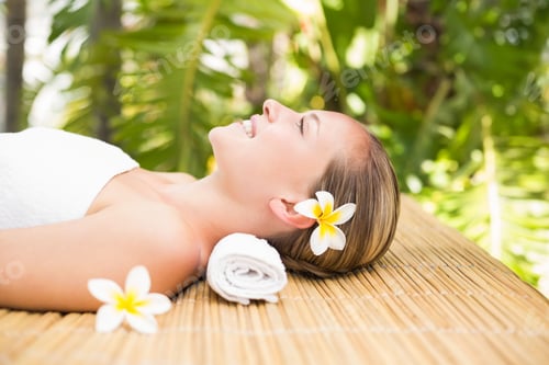 Preview: Woman reclining on bamboo massage table outdoors at spa wrapped in towel with frangipani flower