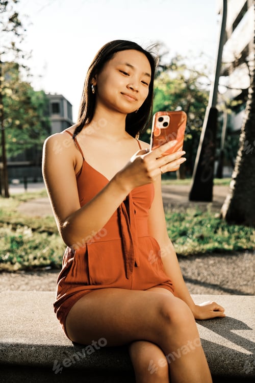 Preview: Young Asian woman using her mobile phone while relaxing sitting on a bench in the park.