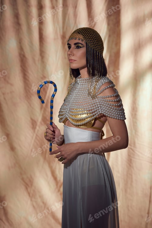 Preview: Stylish woman in egyptian look and pearl top holding crook and standing on abstract background