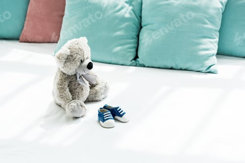 Preview: grey teddy bear near baby sneakers and pillows on white