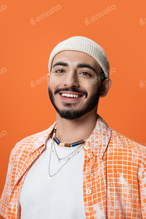 Preview: portrait of stylish bearded man in beanie and beads smiling at camera isolated on orange