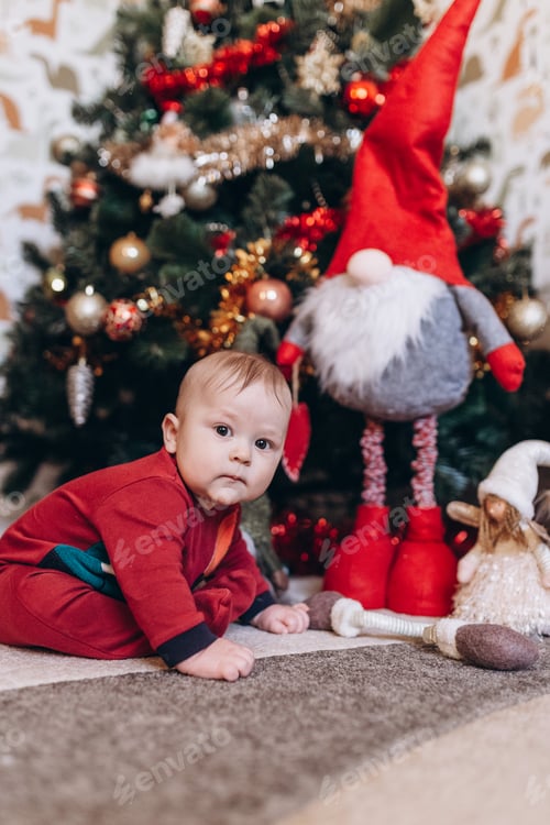 Preview: baby in red Christmas pajamas sits on the background of a Christmas tree with gifts