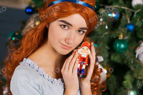 Preview: young woman in a beautiful dress sits near a Christmas tree holding a toy in her hands