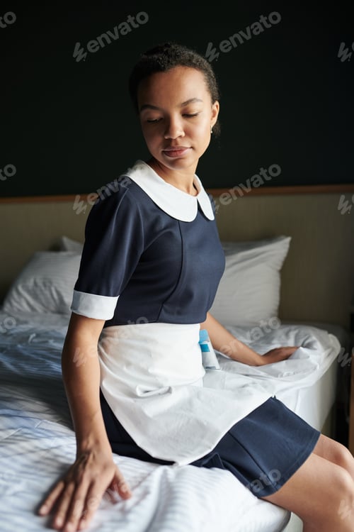 Preview: Young restful housemaid in uniform sitting on clean bed in hotel room