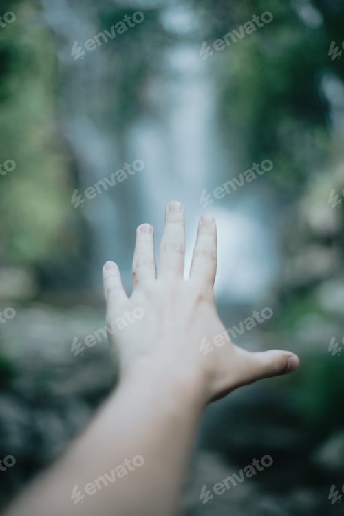 Preview: Moody image of an selective in focus hand in front of a waterfall during a sunny day,