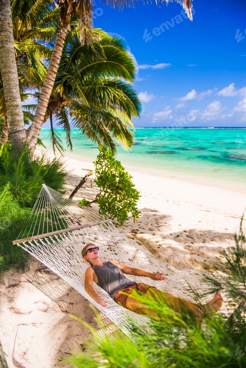 Preview: Woman relaxing in a hammock under palm trees on the white sandy beach on the tropical island of Raro