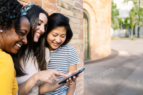 Preview: Three smiling young women using mobile phone together outdoors
