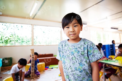 Preview: Young boy standing in a classroom in a Japanese preschool, smiling at camera.