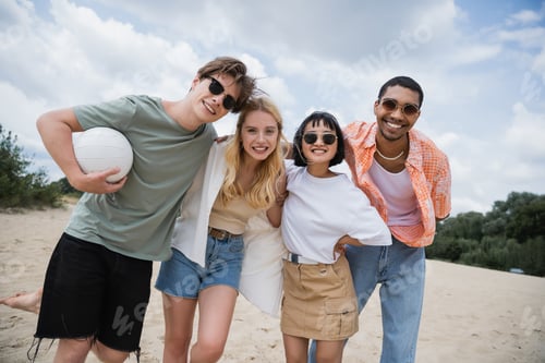 Preview: cheerful multiethnic friends in sunglasses smiling at camera on beach
