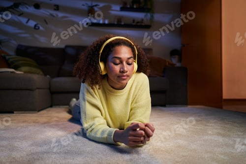 Preview: Young African American girl lying relaxed in living room listening to music wireless headphones.