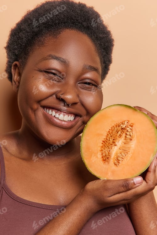 Preview: Vertical shot of dark skinned African woman with curly hair smiles pleasantly holds half of sweet
