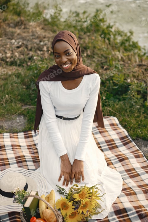Preview: A muslim woman sits on the plaid picnic blanket near the river