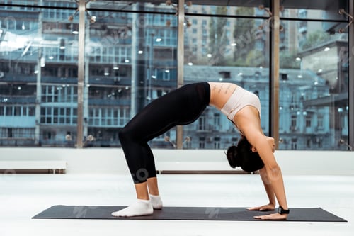 Preview: Sportive girl doing stretching exercises in bridge pose on mat in fitness gym with panoramic windows