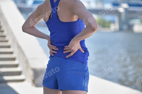Preview: Athletic woman in blue sportswear rubbing the muscles of her lower back, cropped torso portrait