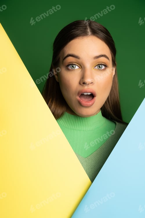 Preview: Shocked teen girl in jumper looking at camera near colorful paper isolated on green