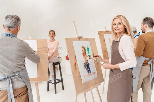 Preview: mature woman smiling at camera while painting on easel at art class