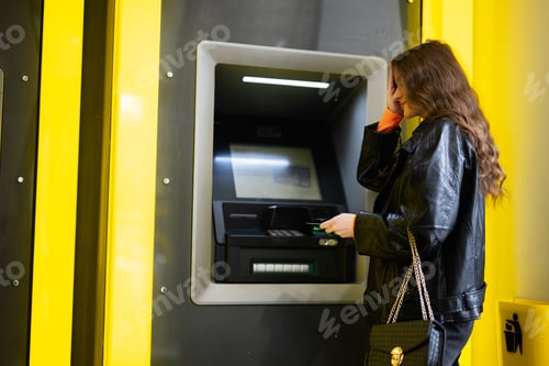 Preview: Young disappointed brunette girl withdrawing money from credit card at yellow ATM.