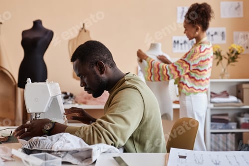 Preview: Side view of young male fashion stylist sewing new items for seasonal collection
