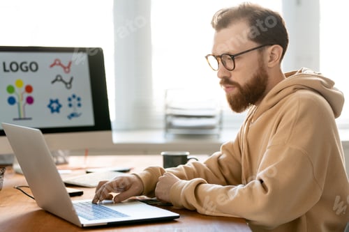 Preview: Young bearded designer looking at laptop display while pressing button on keypad