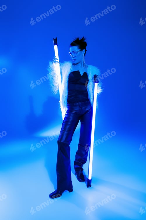 Preview: Stylish woman in furry jacket and sunglasses holding lamps on blue background