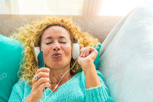 Preview: one happy and young woman singing and listening music on sofa at home with headphones