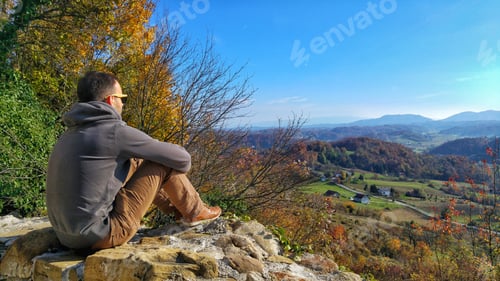Preview: Rear view of Young man sitting on stone wall looking at amazing view of autumn landscape.