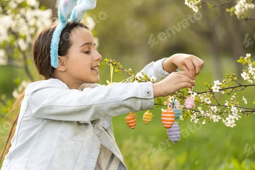 Preview: Adorable little girl in bunny ears, blooming tree branch outdoors on a spring day. Kid having fun on